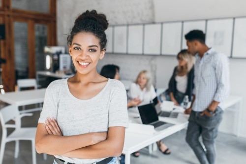 pleased-woman-with-light-brown-skin-posing-with-crossed-arms-smiling-while-people-her-working-indoor-portrait-tired-students-with-laptop-african-curly-girl (1)